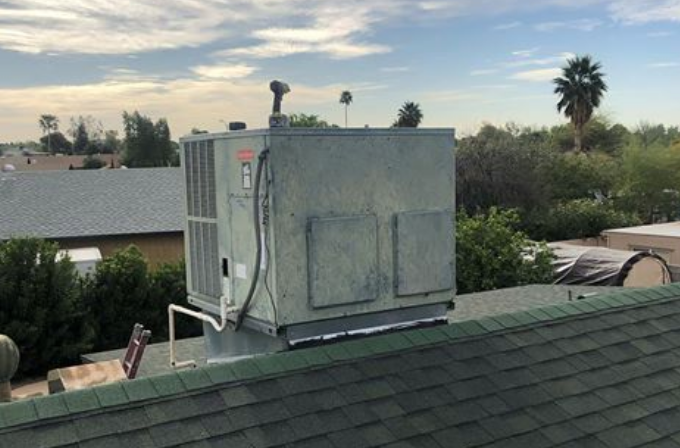 A large, seasoned rooftop HVAC unit installed on a shingled residential roof with a power tool resting on top, showing a neighborhood and palm trees in the background.